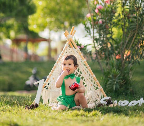 Baby in green outfit sitting in grass next to teepee.