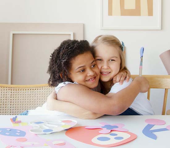 Two girls embracing at a table filled with handmade paper crafts, expressing joy and friendship.